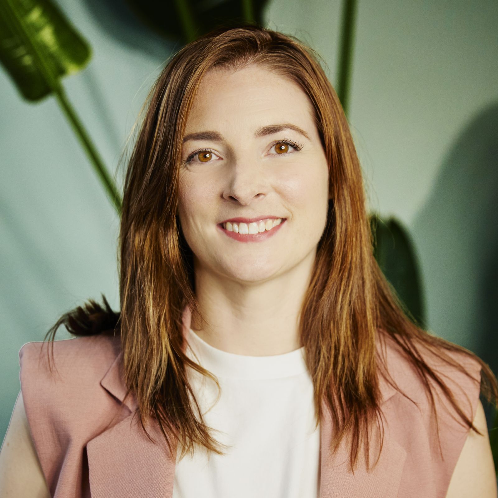 Alice has long red hair, is wearing a pink blazer and is smiling